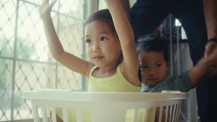 Two children are playing in a laundry basket indoors