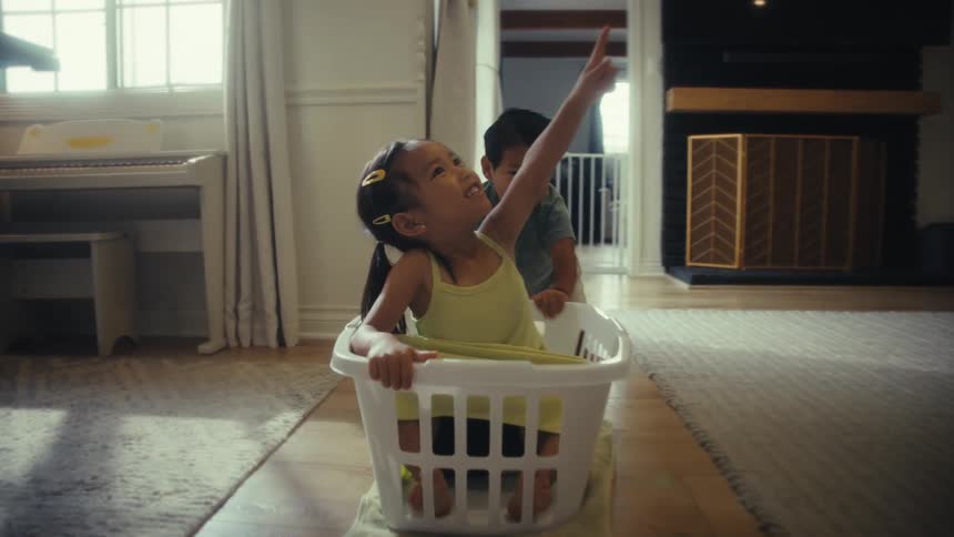 A brother and sister play in a laundry basket in the living room