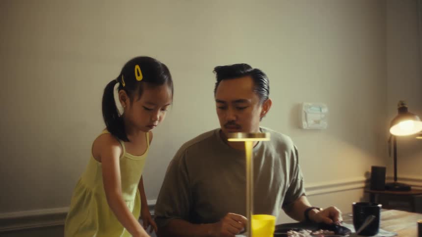 A young girl in a yellow dress stands next to a man at a dining table picking food from a plate and then appears to sneeze or react to something while the man looks on with concern