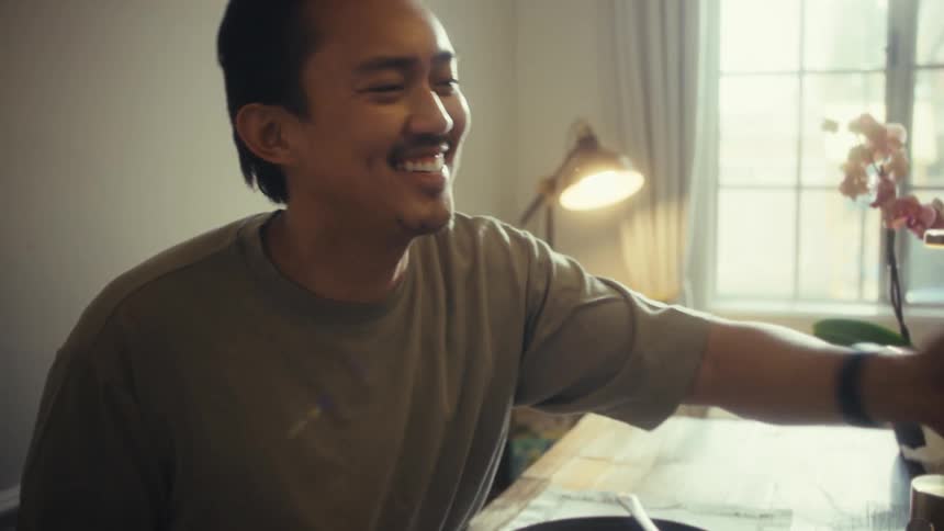 A man holding a mug sits at the dining table smiling as he playfully teases someone seated across from him