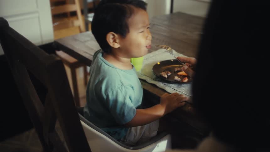 Little boy sitting at the table eating with the help of a person