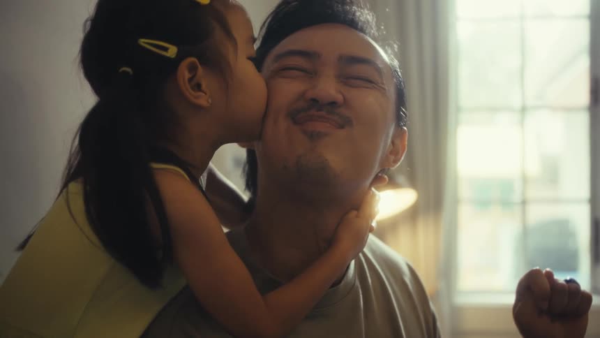 A man is sitting at a table when a little girl comes up from behind and hugs him she then leans in and gives him a kiss on the cheek which makes him smile