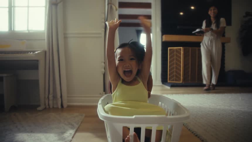 Two children playing with a laundry basket while her mom is on the background in an indoor setting