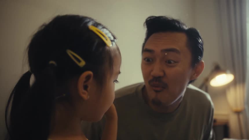 A father and his young daughter are seated at a table where the daughter feeds her father a piece of food and he then leans forward and kisses her