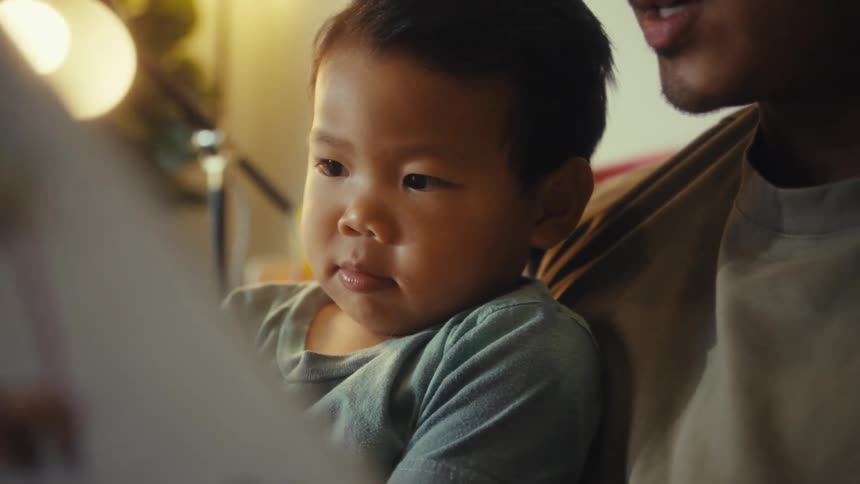 Young adult man sitting on a couch reading a book to a toddler on his lap indoors