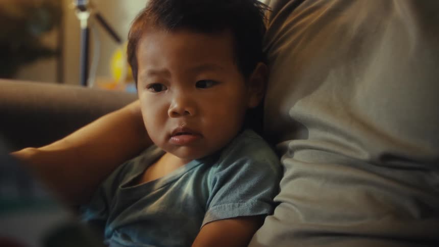 A father holds his young son on his lap as they read a book together in a dimly lit room