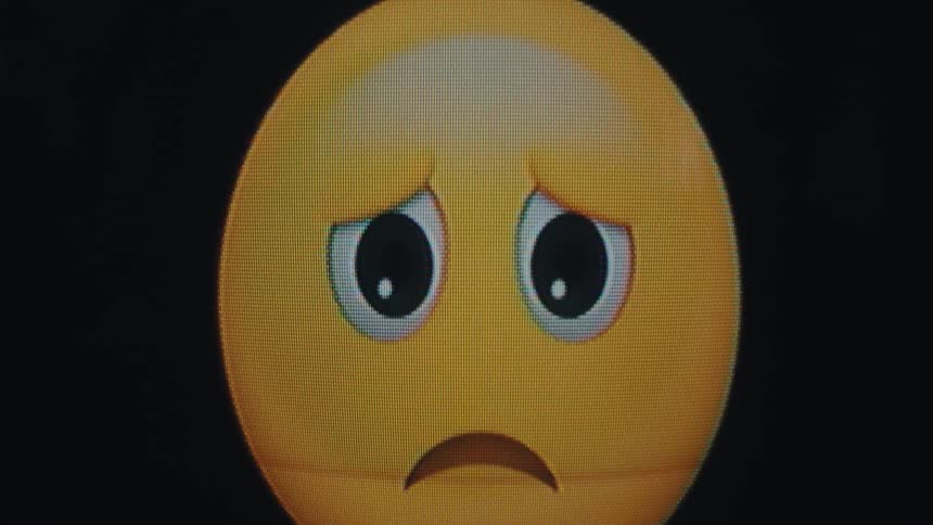 A digital animated sad face emoji with a yellow face downturned brown eyebrows and wide white eyes with black pupils zooms into a close view on a black background