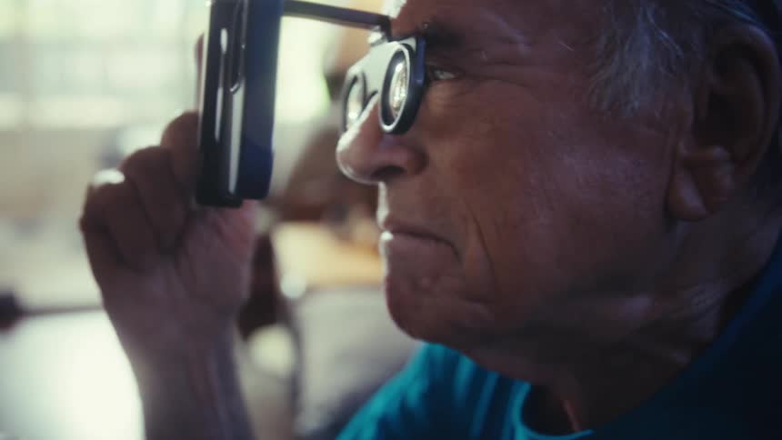 A profile closeup shot of a smiling older man wearing a pair of glasses with an attachment the attachment has two cylindrical lenses that seem to be magnifying a screen