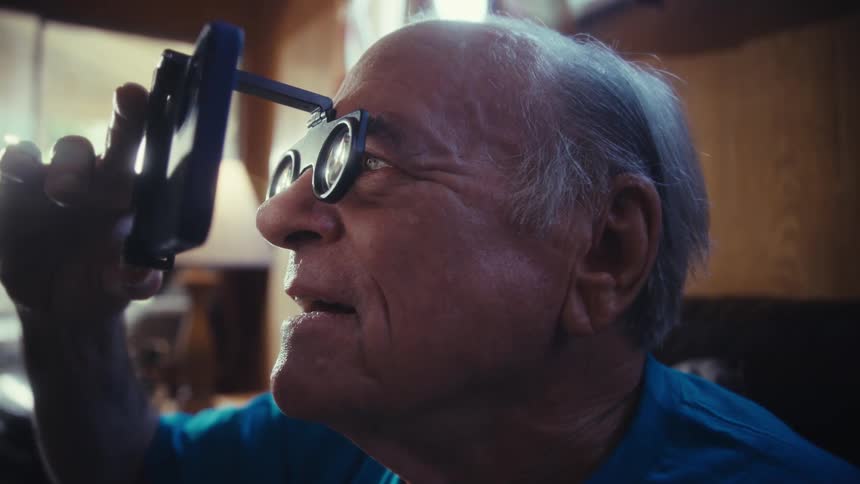 An elderly man with white hair holds a smartphone with a magnifying device attached to it
