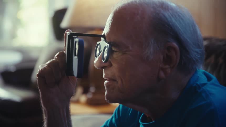 A man with gray hair sits on a couch and looks upwards with a subtle smile while holding a phone with a small magnifying attachment up to his face