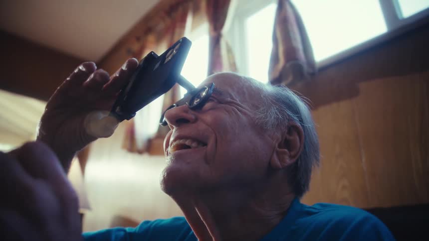 An elderly man wearing goggles holding his phone close to his eyes excitedly watching a program in his living room at midday