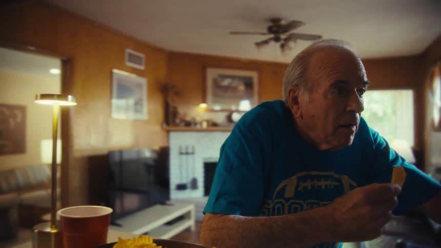 Elderly man wearing a blue football shirt cheering and talking at a television streaming the football game indoors during day time