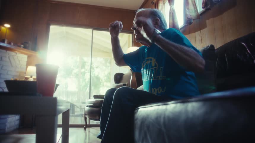 An elderly man in a blue tshirt sitting on a couch in a sunlit living room reacts with excitement to something hes watching off screen suddenly shouting and gesturing emphatically with his arm