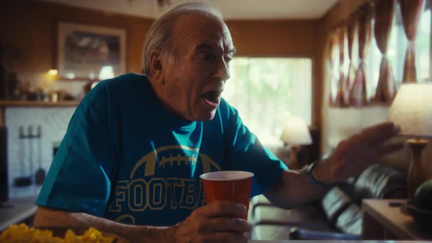 A person holds a cup and gestures with a hand while looking at a television screen and reacting with a facial expression