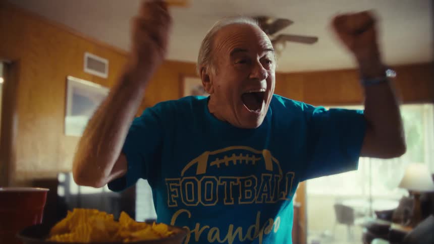 An elderly man wearing a football grandpa t shirt enthusiastically cheers while watching a screen and eating potato chips at his kitchen counter