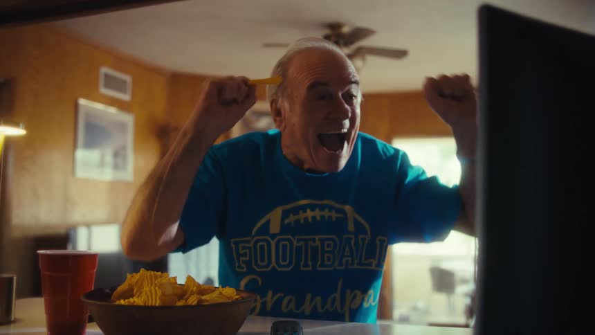 Elderly white man wearing a football shirt cheering in front of a monitor over a counter next to a bowl of nachos and a cup indoors during day time
