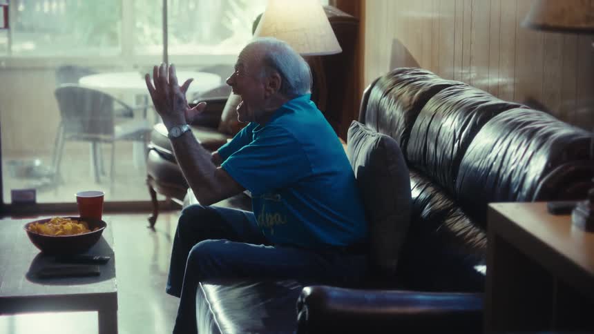 An older male sits on a couch enthusiastically gesturing toward a television