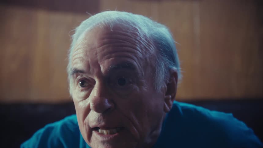 The video shows a man with white hair wearing a blue tshirt with the words football grandpa sitting on a leather couch he is intently watching something offscreen with his expression changing from anticipation and surprise to a smile
