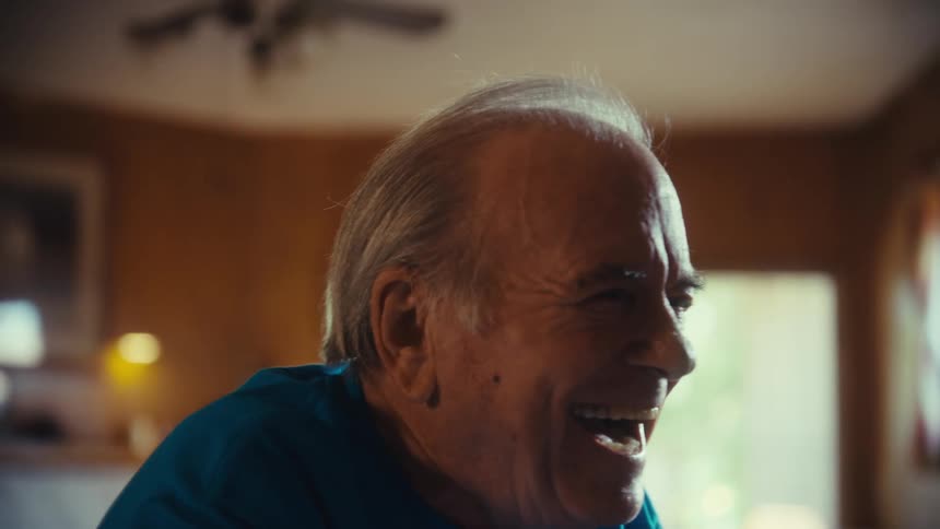 A video shows a closeup of an elderly man with gray hair and a blue tshirt he seems to be watching something and then starts to laugh heartily taking a drink from a red cup between laughs