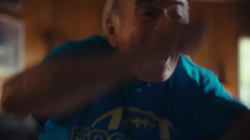 An elderly man in a blue shirt is sitting in a living room eating snacks and watching tv he suddenly gets excited smiles and starts cheering enthusiastically with a snack still in his hand