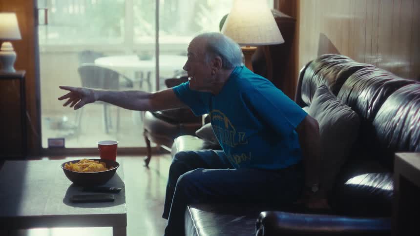 An elderly man sits on a couch in his living room looking excited while watching a game at midday