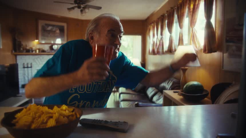 A person sits at a counter in a living room looks at a television and reacts with movement and a facial expression while eating