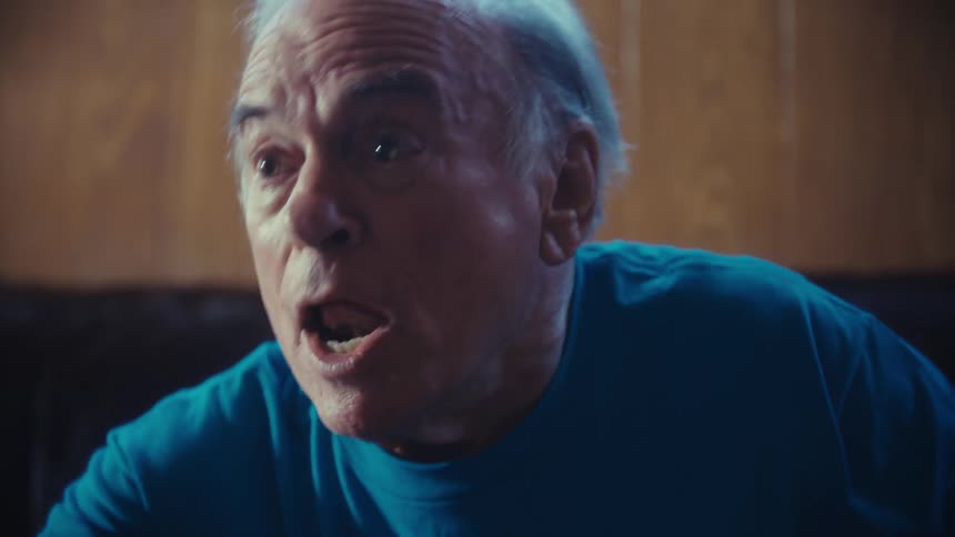The video shows an older man with graying hair sitting on a brown leather couch in a dimly lit room he is wearing a blue tshirt with football grandpa written on it and is watching a game excitedly throwing his hands up in the air