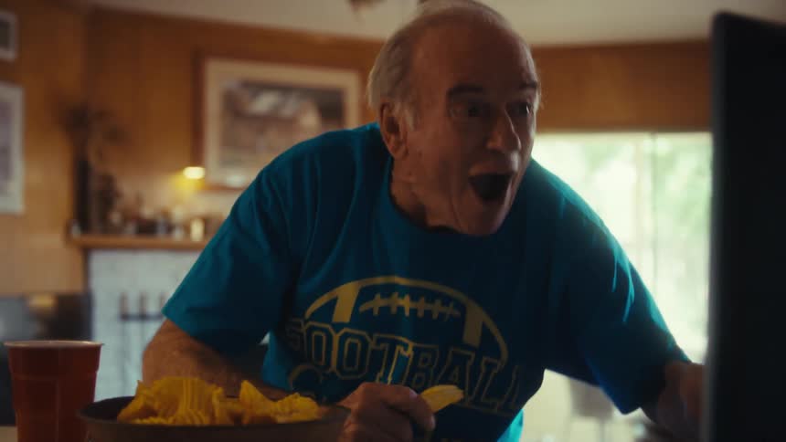 A happy man in a blue shirt with a football graphic eats a chip from a bowl and reacts with a loud cheer and a closed mouth laugh as he watches something on a computer monitor