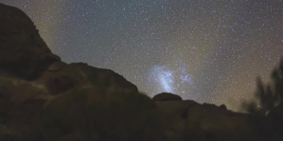 The night sky is filled with a multitude of stars and a formation of stars above a terrain with rocks and some foliage