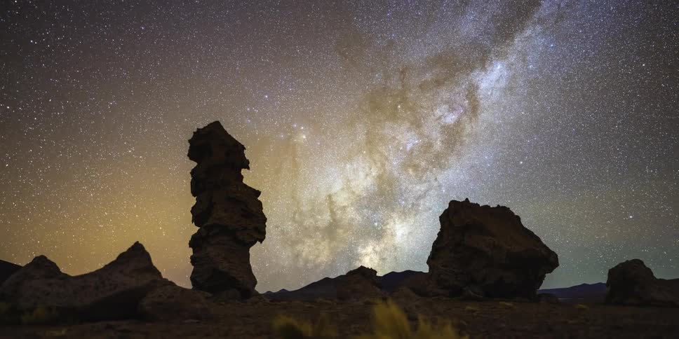 A time lapse of a starry night sky shows the milky way moving behind silhouetted rock formations with a warm glow on one horizon