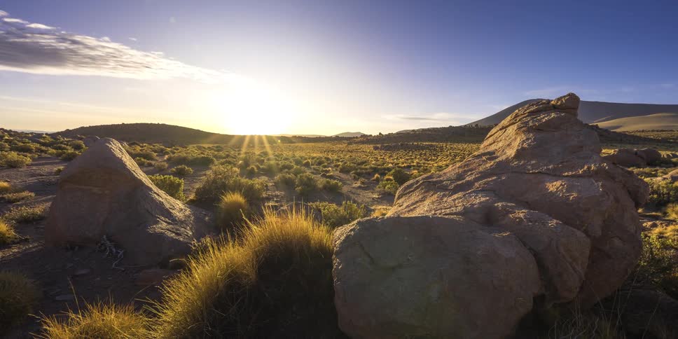 The sun sets behind a distant mountain range casting warm bright rays of light over a vast rocky desert landscape filled with small bushes and large boulders