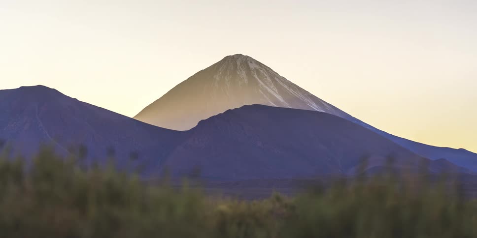 A tall mountain with snow on its peak stands in a landscape with other mountains under a sky with a sun