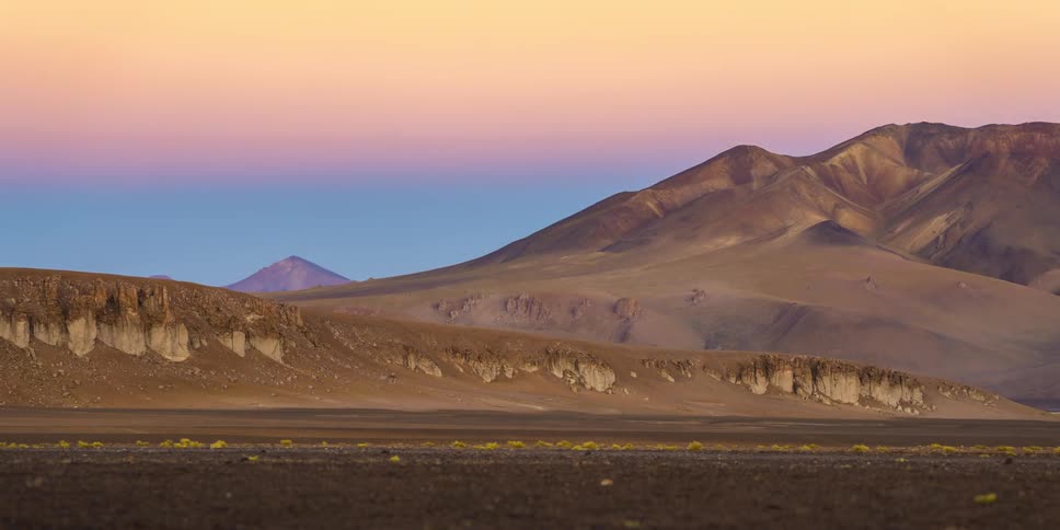 A timelapse video shows a golden desert landscape with mountains in the background the sun sets casting long shadows across the ground and changing the color of the sky from a light blue and yellow to a deeper orange and purple hue