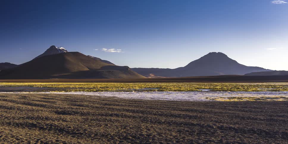 Clouds drift over a vast arid landscape with a snowcapped mountain a marshy area and distant peaks as the scene transitions from a bright blue sky to a warm golden sunset