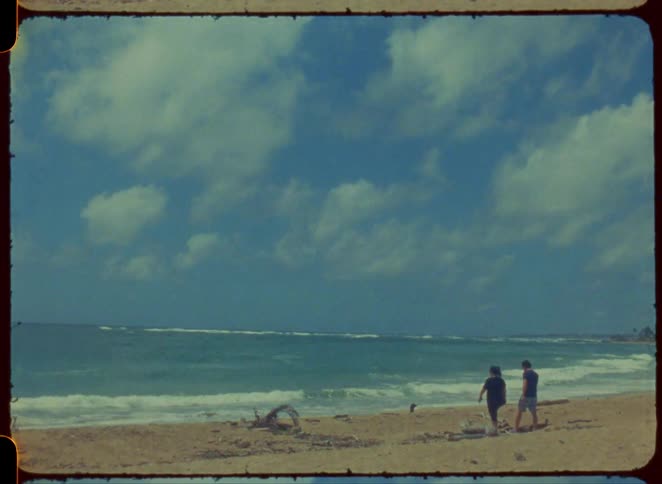 Two people walk on a stretch of sand with waves of water moving on the shore under a sky with clouds