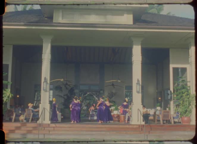 Dancers in folk costume performing to an audience at the porch of an old house outdoors during day time