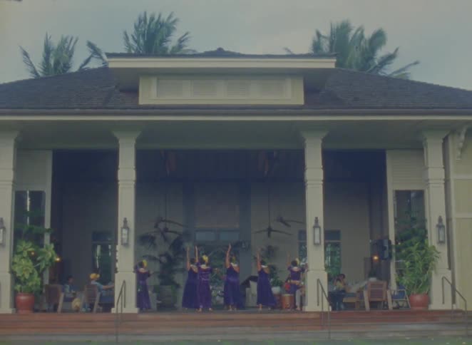 A group of people wearing similar outfits dance on a lawn in front of a building with columns and a veranda