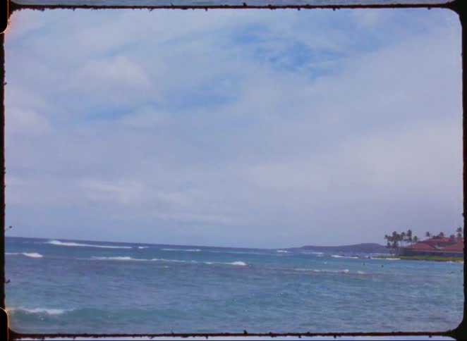 A body of water with waves moves towards a landmass with trees under a sky with clouds