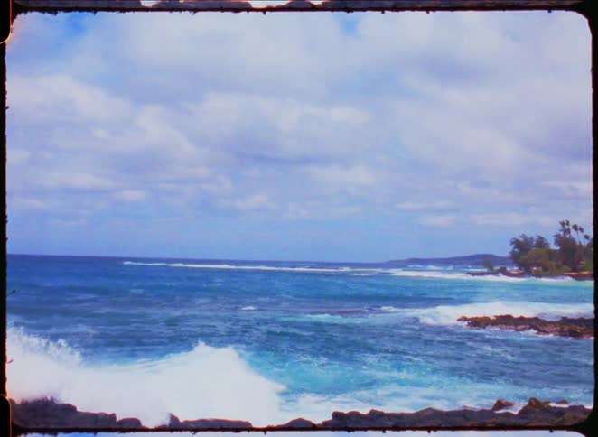 A blue ocean with rolling white waves breaks on a dark rocky coastline with green bushes and trees under a cloudy sky