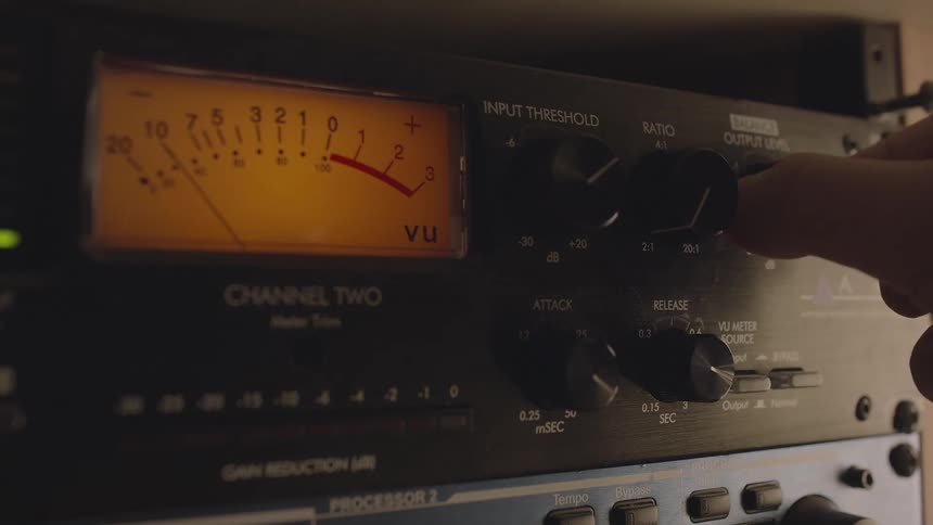 A hand adjusts the output level knob on a piece of audio equipment while a vintage style vu meter shows the signal level and other controls are visible on the panel