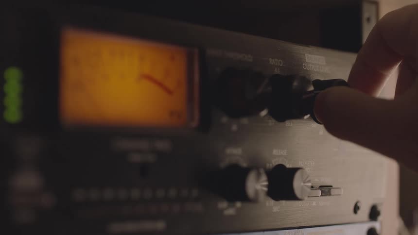 A hand adjusts a knob on a piece of audio equipment with an analog meter while various other controls and indicator lights are visible on the panel
