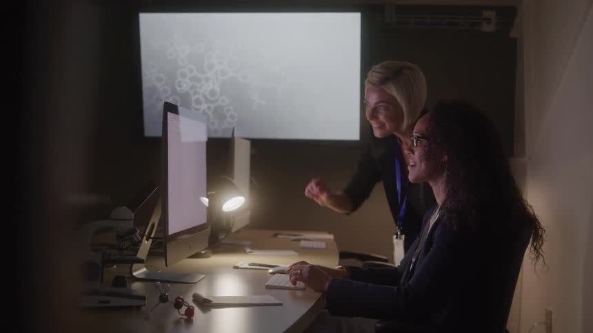 A blond woman in a black shirt stands over a seated woman with long dark hair and they both look at a computer screen and smile as the blond woman points to the screen