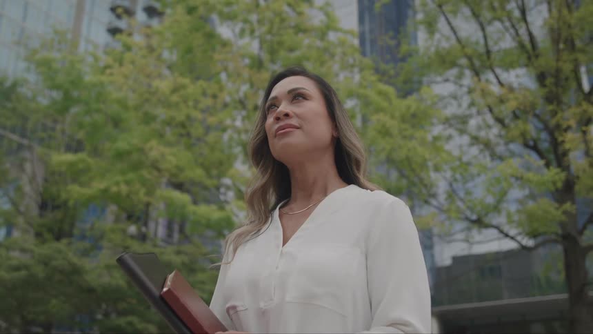 A woman wearing a white shirt stands outside a city building holding two notebooks at midday