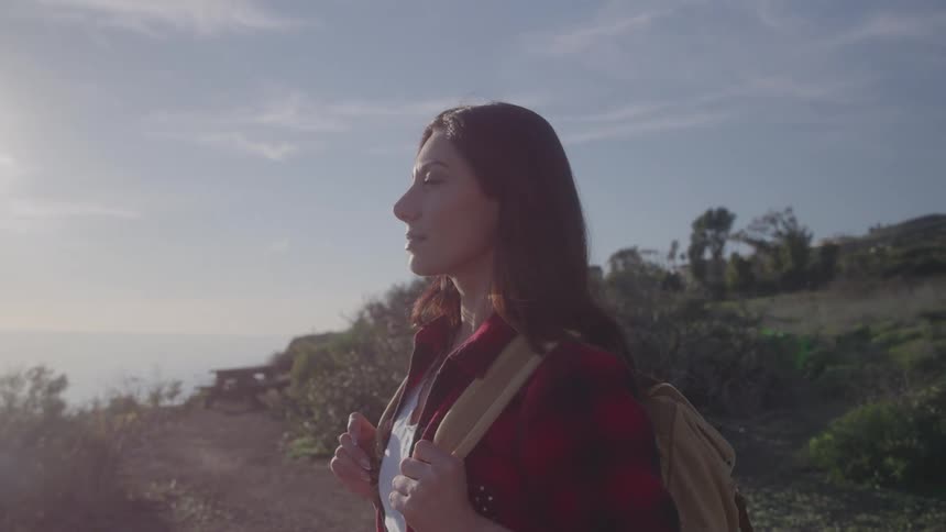 A woman with a backpack stands on a coastal trail gazing at the ocean