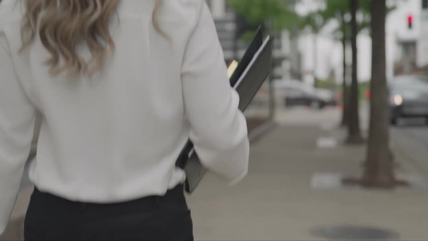 A woman in a white blouse and black pants walks down a city sidewalk holding a binder under her arm as the camera follows closely behind her