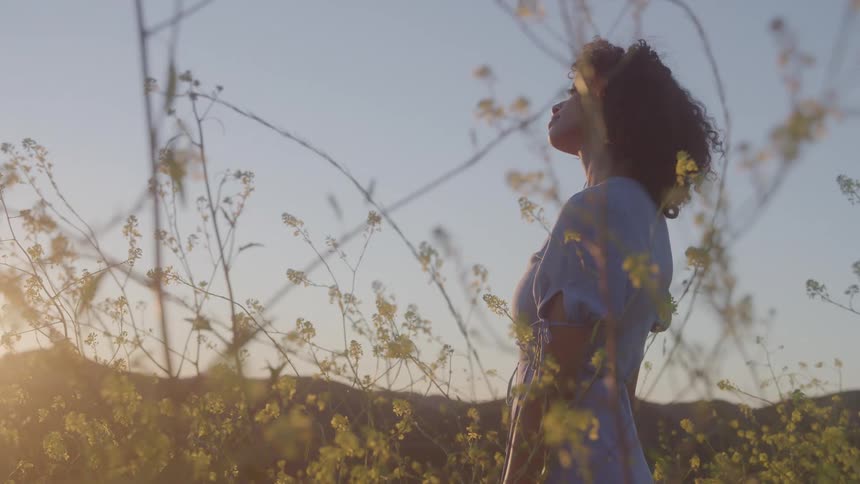A woman with long curly hair stands in a field of yellow flowers and green plants looking up with her head tilted to the side as the sun sets behind her