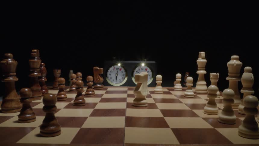 A chessboard set up for a game with two people playing in a dark room and two chess clocks in the background