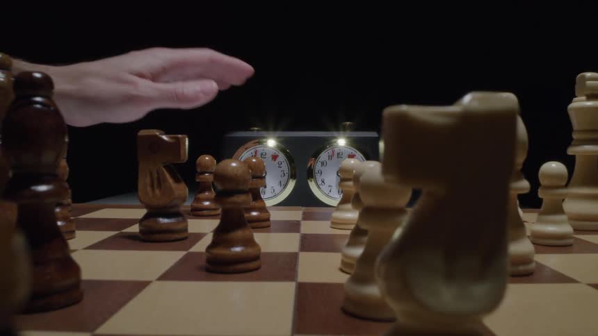 Two hands moves pieces on a chess board as a clock sits on a table
