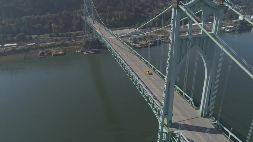 An aerial shot captures a bright yellow car driving across a large light blue suspension bridge that spans a wide river with an industrial shoreline in the background