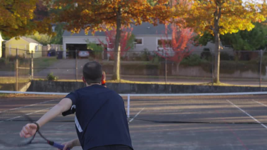 Young man and young woman playing tennis on a tennis court next to a suburban neighborhood during dusk or dawn